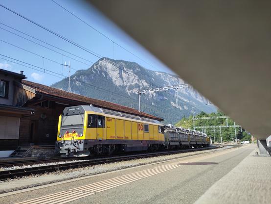 Foto. Eine gelbe Diesellokomotive mit Schotterwagen im Bahnhof.