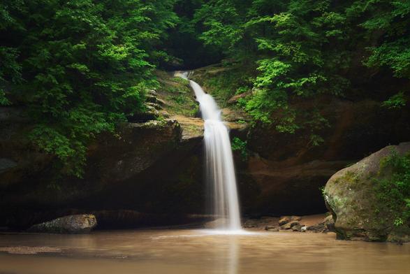 A narrow waterfall streams over mossy rock into a calm, muddy pool, framed by lush green forest and large boulders.