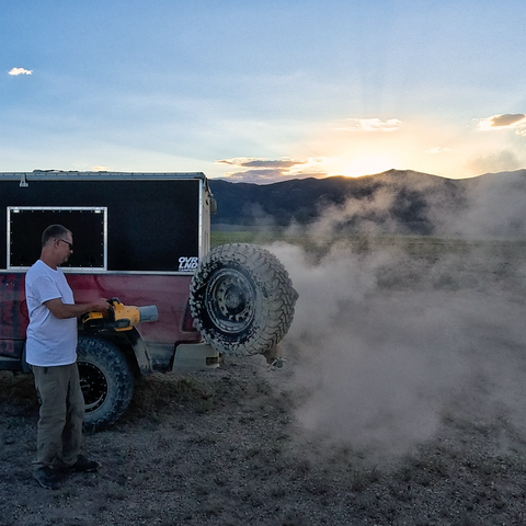 A man uses a handheld blower to clear dust from a spare tire mounted on the back of a red off-road truck camper at sunset, with a cloud of dust drifting into the open landscape and mountains in the background.