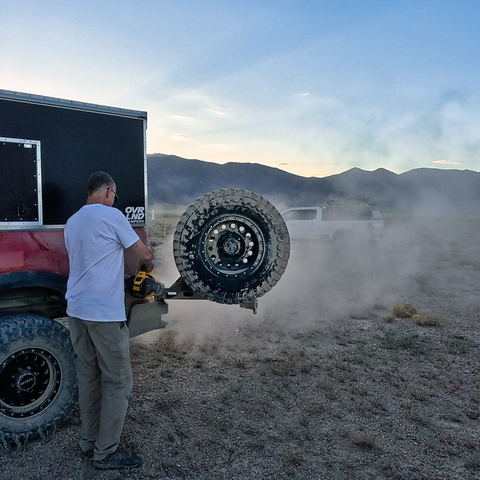 A man uses a handheld blower to remove dust from a spare tire mounted on the back of a red off-road truck camper, with a cloud of dust drifting across the gravel landscape and mountains in the background.