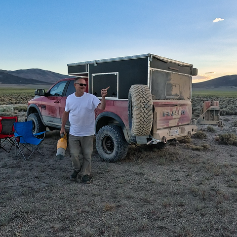 A man in a white t-shirt and sunglasses stands beside a dusty red RAM off-road truck camper in a desert landscape, holding a handheld blower and gesturing with his other hand, with camping chairs and mountains in the background at sunset.