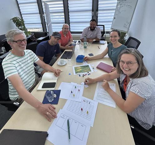 Sechs Personen sitzen um einen Konferenztisch mit verschiedenen Papierausdrucken und bunten Spielfiguren.