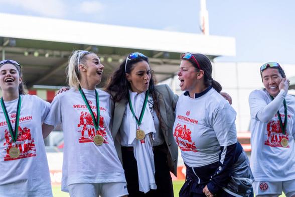 Fünf Mainzerinnen mit "Meisterinnen"-Shirts, Medaillen und Ski-Brillen schunkeln singend und lachend in einer Reihe: Von links nach rechts Vital Kats, Chiara Bouziane, Ebru Uzungueney, besonders inbrünstig feiernd Mentalcoach Sandra Kärger und ganz rechts Mamiko Matsumoto, die sich lachend ein Ohr zuhält.