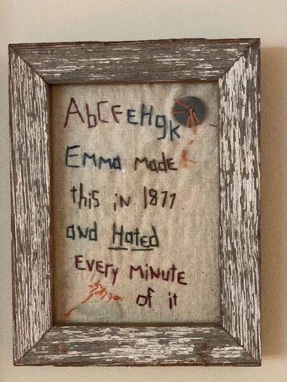A framed piece of embroidery. The wooden frame looks old and splintery. It begins with a badly-sewn alphabet from a to k, then says, Emma made this in 1877 and hated every minute of it.
