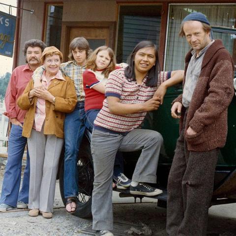 A vintage promotional photo from the Canadian drama series 'The Beachcombers.' The image features the main cast members, including Nick Adonidas (right), posing in front of a shop in Gibsons, British Columbia. The scene captures the show's depiction of coastal life and log salvaging in Canada during the 1970s