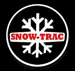 Un logo o emblema circular con un fondo negro. En el centro, un gran copo de nieve estilizado de color blanco. Sobre el copo de nieve, una banda horizontal blanca con las esquinas redondeadas contiene el texto "SNOW-TRAC" en letras mayúsculas rojas y negritas. Todo el diseño está enmarcado por un borde circular blanco.