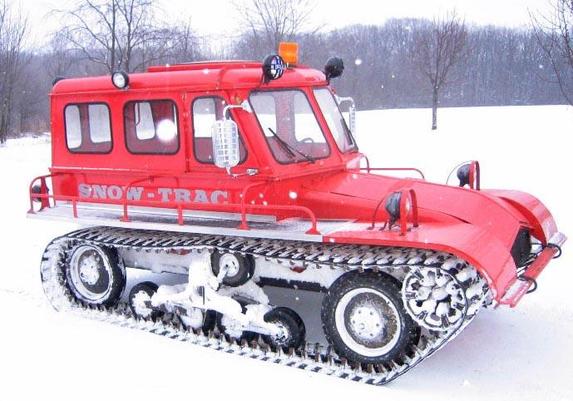 Una imagen muestra un vehículo oruga (snowcat) de color rojo brillante, identificado con la inscripción "SNOW-TRAC" en su costado. Está estacionado en un paisaje invernal completamente cubierto de nieve fresca. El vehículo tiene una cabina rectangular con varias ventanas y un parabrisas inclinado. En el techo de la cabina se observa una luz de advertencia naranja y dos faros redondos negros. La parte delantera del vehículo es aerodinámica e inclinada, con dos faros adicionales. A lo largo de los costados, debajo de las ventanas, hay barandillas de seguridad metálicas. Sus anchas orugas dentadas, diseñadas para la nieve, están parcialmente cubiertas de blanco, dejando ver las ruedas y el mecanismo interno. El fondo de la imagen presenta un campo nevado con varios árboles sin hojas, indicando un entorno forestal o rural en invierno.