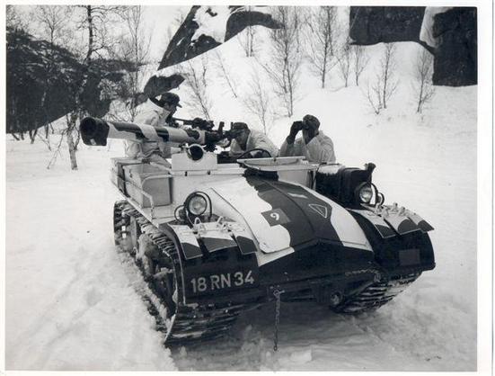 Una fotografía en blanco y negro muestra un vehículo militar blindado sobre orugas, con un patrón de camuflaje invernal blanco y negro, avanzando por un paisaje cubierto de nieve. En la parte trasera del vehículo está montado un cañón o lanzador antitanque. Tres soldados con uniformes de camuflaje blanco están en el vehículo: uno opera el cañón, otro observa el horizonte con binoculares, y un tercero está al volante. El vehículo lleva la matrícula '18 RN 34' en la parte delantera y un número '9' con un triángulo en el capó. El fondo muestra árboles desnudos y formaciones rocosas cubiertas de nieve, indicando un ambiente invernal.
