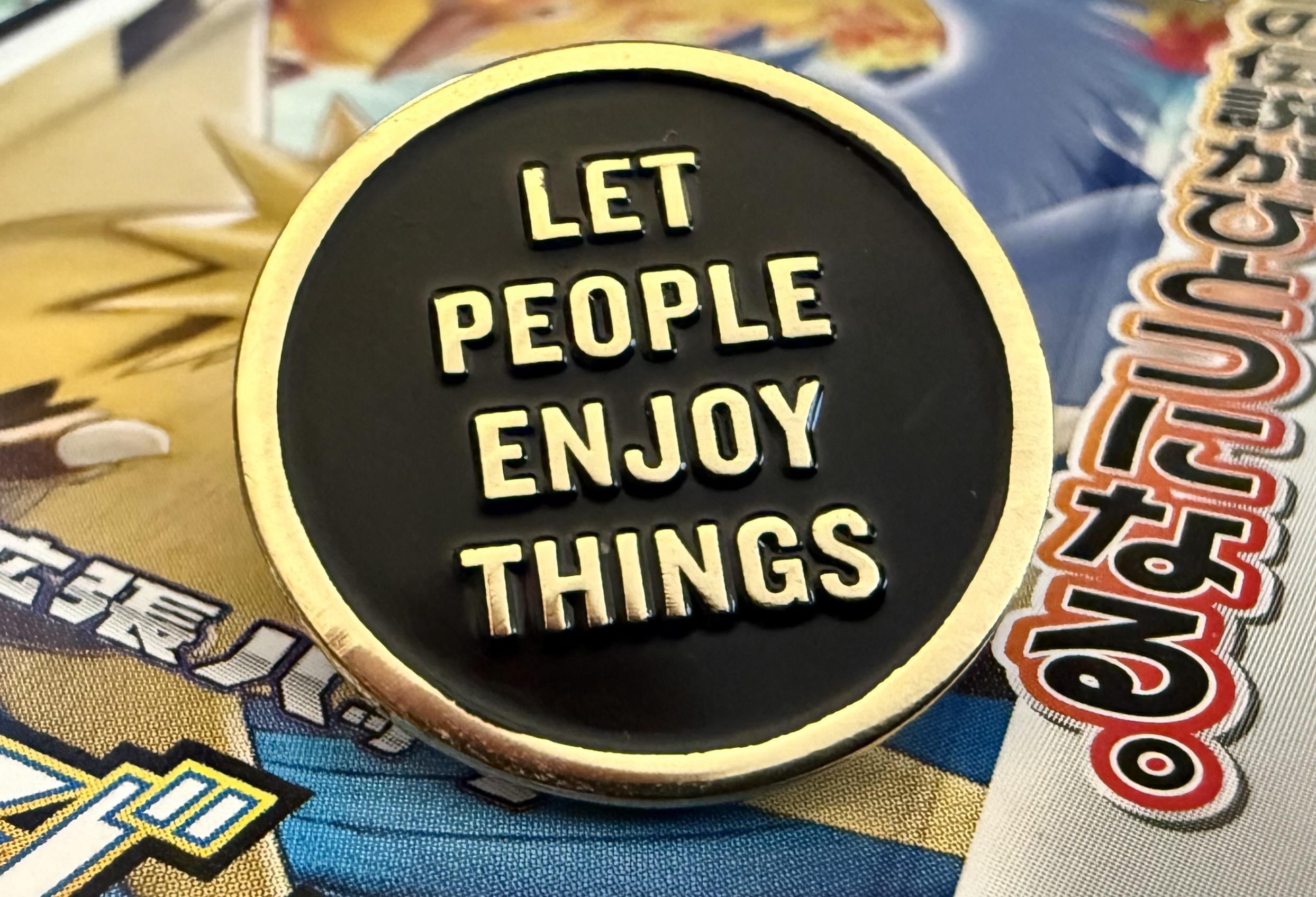 An enamel pin that says “LET PEOPLE ENJOY THINGS”, in gold text against a round black circular background with a gold edge around the circle.
