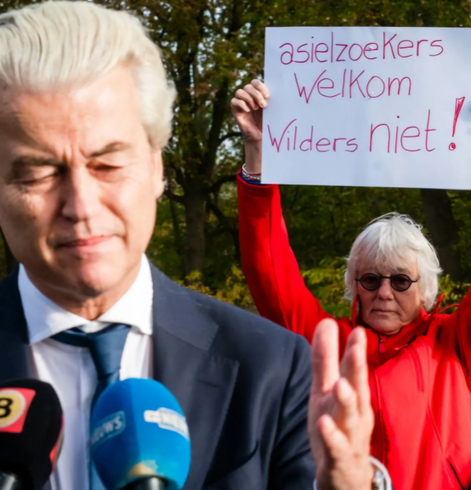 Foto van Geert Wilders met achter hem een persoon die een bord omhoog houdt waar "asielzoekers welkom, Wilders niet" op staat