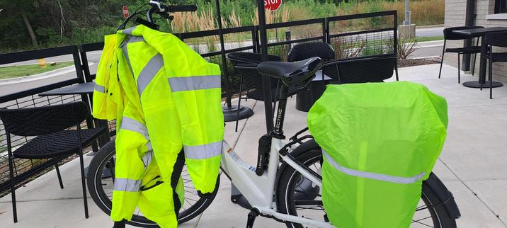 The image shows a white electric bicycle parked on a concrete patio. The bicycle is equipped with a bright neon green rain cover on the rear rack and a matching neon green jacket with reflective stripes hanging on the handlebars. The jacket is designed for high visibility, featuring reflective stripes on the sleeves and torso. The bicycle is positioned in front of a black metal railing, with black metal chairs and tables visible in the background.