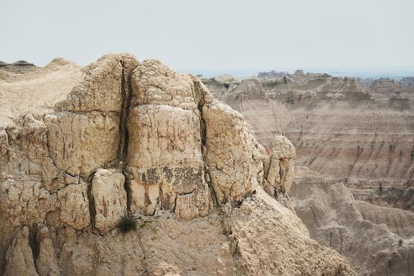 Close-up of rugged, beige badlands rock formations with deep cracks and layers, overlooking a vast eroded canyon under a pale sky.
