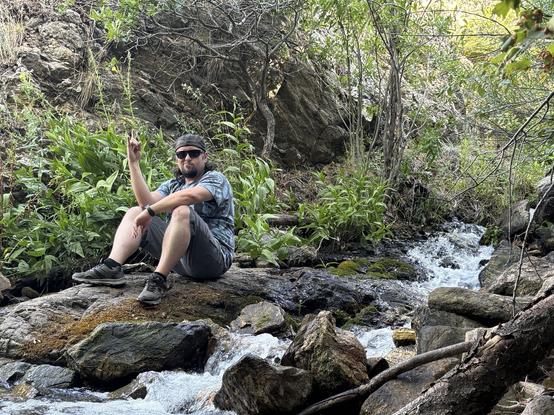 A man with shaggy hair and a goatee, wearing sunglasses, a patterned shirt, shorts, and a backwards baseball cap sits on a mossy boulder in a small, rushing creek in a lush, rocky forest. His legs are bent and he is resting his arms on his knees as he makes a devil horn gesture with his right hand while looking toward the camera. Green plants grow densely along the water, and the scene is framed by rocks, branches, and trees.