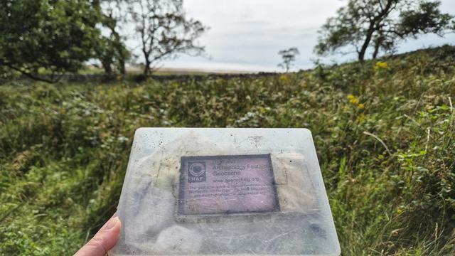 Das Bild zeigt eine Hand, die einen durchsichtigen Plastikbehälter hält. Auf dem Behälter ist ein Etikett mit der Aufschrift „Highland Archaeology Festival Geocache“ zu sehen.  Der Hintergrund besteht aus einer unscharf dargestellten Landschaft mit Gras, Büschen und Bäumen, die auf eine ländliche Umgebung hindeuten. Der Fokus liegt auf dem Behälter, der wie ein Fundstück oder ein Teil eines Geocaching-Spiels aussieht.