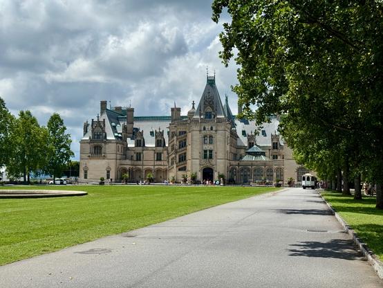 A large house that resembles a castle with an elaborate design of stone brick facade, spires, beautiful windows, and beautiful grounds. There is a huge green lawn stretching out in front of it with an access road circling around the lawn. Trees line the edges of the access road. There are puffy clouds in the sky, but the sun was shining for this moment. Tour buses and people are also seen on the grounds.