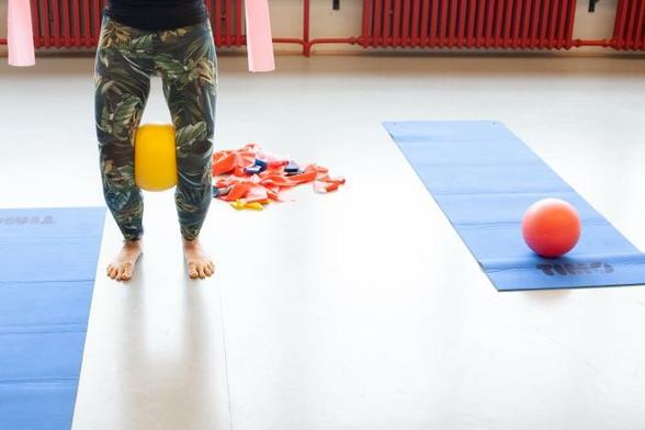 Person in gemusterten Leggings macht Sportübungen mit einem gelben Ball zwischen den Beinen, daneben sind Yogamatten und eine orangene Kugel. Foto: Sandra Ratkovic