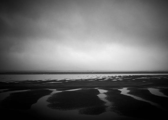 A black and white photograph of a misty estuary. The foreground shows dark, rippling water patterns with patches of exposed land. Beyond that lies a flat, distant horizon under a cloudy sky, creating a calm and tranquil mood