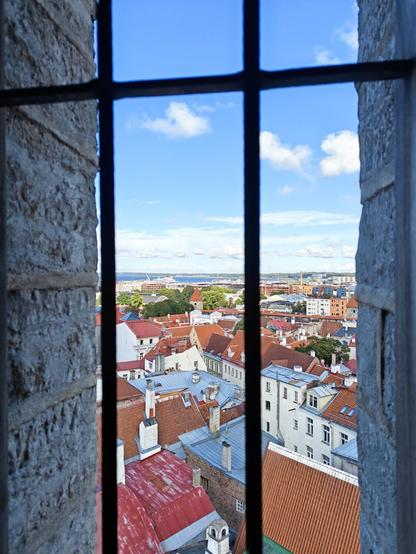 Picture of the old town of Tallinn taken from above the rooftops in a tower. The view includes lots of old white buildings with red rooftops. The sky is blue with some white clouds. In the foreground you can see two vertical and one horizontal bars, that are attached to the window hole for safety reasons.