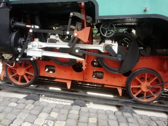 Close-up side view of an antique steam train chassis showcasing the intricate mechanics; visible components include large orange wheels, connecting rods, a black driving wheel, and a red and black framework. The train rests on a metal track secured by a chain, with a cobblestone surface in the foreground.

Nahaufnahme der Seitenansicht eines antiken Dampflokomotiv-Fahrgestells, das die komplexe Mechanik zeigt; sichtbare Komponenten sind große orangefarbene Räder, Pleuelstangen, ein schwarzes Antriebsrad und ein rot-schwarzes Gestell. Die Lokomotive steht auf einer mit einer Kette gesicherten Metallschiene, im Vordergrund ist eine Kopfsteinpflasteroberfläche zu sehen.