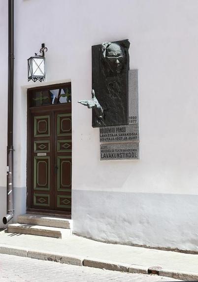 The outside of a building in a street, a drainpipe runs usefully but irrelevantly down the left, debouching I think onto the pavement. To the right, a black panelled door is up two steps, a lantern above which may be of less utility if this text is useful.
To the right a sculpture beseeches in high relief from the wall, with an account of history here in IIRC a past occupation. There is very little colour.