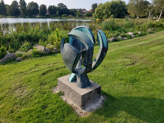 A sculpture at the Haderslev Dam. It's round-ish and cut into three slices which doesn't look as if they fit together because the slices have all different forms and pieces. It's a green grey color and is placed on a small pedestal.