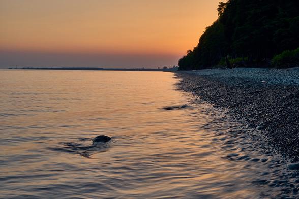 A tranquil shoreline at sunset, featuring calm waters and a pebbly beach. The sun casts warm hues across the sky and reflects on the water, with trees lining the coast in the background.