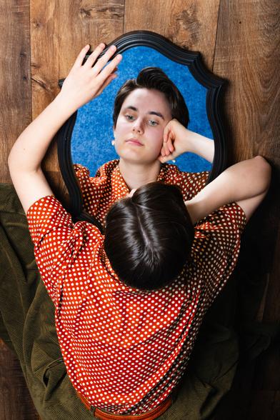 The image shows a person with short hair looking at their reflection in an ornate mirror on a wooden floor. They are wearing a red polka dot shirt and resting their chin on one hand. The angle creates a unique perspective where we see both the back of their head and their face in the mirror. The reflection shows a calm, introspective expression with a blue background surrounding the mirror.