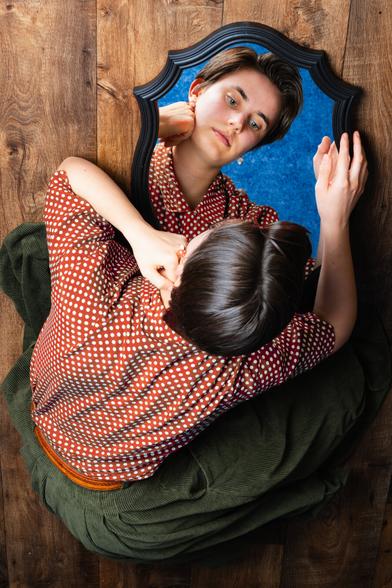 The image shows a person with short hair seated on a wooden floor, looking into an ornate mirror. They are wearing a red polka dot shirt and green pants. The person rests their chin on one hand, with the other hand touching the mirror’s edge. In the mirror, their face reflects a calm, introspective expression against a blue background.