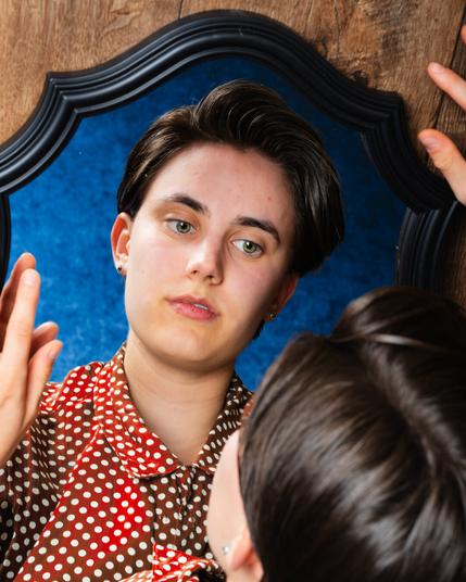 The image shows a person with short, dark hair gazing at their reflection in an ornate mirror. They are wearing a red polka dot shirt. The close-up perspective captures their calm expression as they rest their hand on the side of the mirror. The background features a blue textured surface, framed by wooden planks.