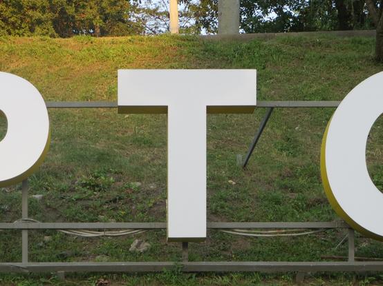 Белая буква Т, установленная на металлической конструкции на придорожном газоне. На краях кадра частично видны соседние буквы. // A white T mounted on a metal structure on a roadside lawn. The adjacent letters are partially visible at the edges of the frame.