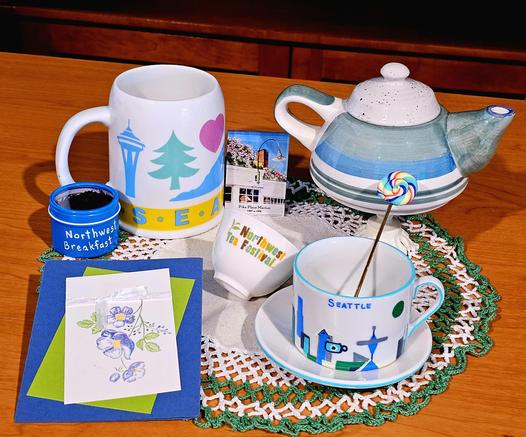 Tea tableau in green, white, and blue on a white crocheted doily with a bright green ruffle. There are a handpainted cup and saucer with a motif of Seattle landmarks, a large mug with similar designs, an earthenware teapot on a pedestal, a drawing of blue flowers, a gaiwan cup with the logo Northwest Tea Festival, a miniature painting of Pike Place Market, and Northwest Breakfast black loose-leaf tea from Market Spice in Seattle.
