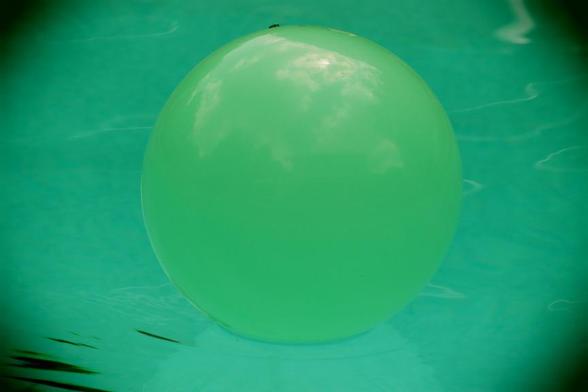 A surreal, dreamy close-up of a yellow beach ball floating on the surface of the water in a swimming pool is in a green toned retro-colour.  Resting on the top of the ball is a black ant, wondering how it got there, and if the cold medication it took is way too strong.