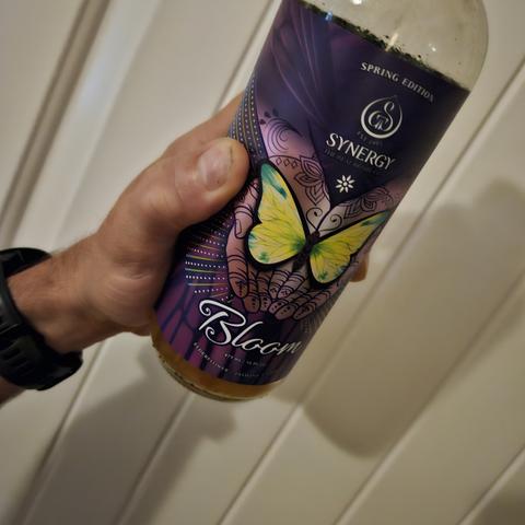 a kombucha bottle by Synergy called Bloom. it is a spring addition and features dark purple hands holding a yellow green winged butterfly, that when looked at from afar, looks like spiderman in the dark.