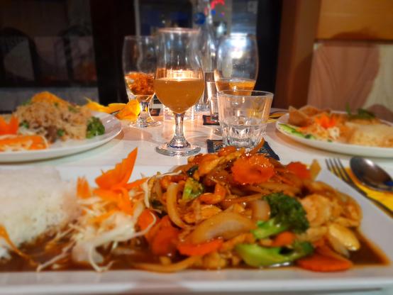 Focus is on a thai-style stir-fry and rice on a longitudinal plate. It is sitting in a thin brown sauce and the stir-fry includes cabbage, carrot, brocolli, chicken, and many other seasonal winter vegetables. Two other plates can be seen, with drinks in the middle of the table.