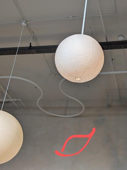 snapshot of a ceiling with two round white paper pendant lights (eyes) and a curving white pipe (nose). i've drawn a mouth for this face.