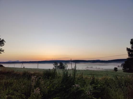 Un lever de soleil. Le ciel rose à bleu à l'horizon. Des montagnes dans le fond, sur lesquelles on distingue à droite des éoliennes. Le tout sous une mer de nuages. Au premier plan, un pré très vert.