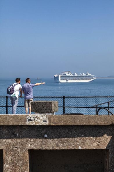 A portrait orientation photo of two men standing on a concrete wall with the sea beyond them. A barrier of rounded black metal with chain fencing between its poles reaches to waist height on the two men. They are facing away from us and standing on the left side of the wall. The man on the left has light jeans and a white, short-sleeved shirt with a purple backpack, and he's leaning forward slightly to rest on the rail. His friend to the right has light brown shorts and a blue short-sleeved shirt but he's standing upright with his right arm pointing straight out to the right, directing the other man's attention to something out of sight. Out to sea in the distance there's a cruise ship. It's not the thing that the man is pointing at but it almost looks like it as his finger nearly touches the white ship's aft from our perspective. The sky is clear blue but hazy with heat and the sea is calm. Bright, hot sunshine bakes the scene.