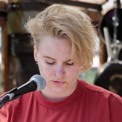 Een foto van Sjoukje Kamphorst met roodbruin t-shirt en hier blond kort haar, die voor een microfoon staat en naar beneden kijkt.