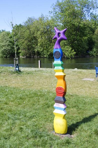 A post in the form of a totem pole, in all the colours of the rainbow on the top. The bottom part has the colours of the transgender flag. It sits in a field, with the river Thames behind it.