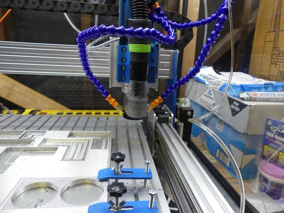 A CNC machine with a focus on the cutting head and the workpiece. The cutting head is equipped with two adjustable blue coolant / lubrication hoses, which is coiled around the head. The aluminum workpiece, secured by blue clamps, features circular and rectangular cutouts. The machine is mounted on a metal frame with linear guides, allowing for precise movement along the X and Y axes. In the background, there are boxes. The setting appears to be a workshop or a manufacturing environment, with various tools and materials visible.