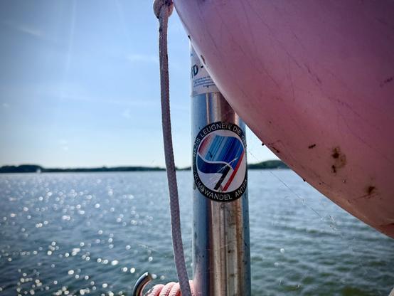 Nahaufnahme eines Metallpfostens AntiFa-Sticker, der teilweise von einem verblichenem Rettungsring verdeckt, im Hintergrund blauer Himmel & die Schlei
