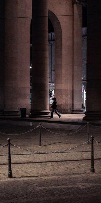 A young woman walks beneath a tall colonnade at night. City lights come from behind the columns.