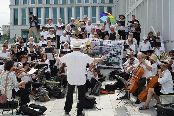 Das Foto zeigt die "Lebenslaute" um das Transparent zur Aktion aufegreiht. Der Text des mit einem Foto eines NATOdrahtzaunes illustrierten Transparentes lautet: "Lebenslaute. Klassische Musik - politische aktion. Mit Pauken und Trompeten gegen Grenzzäune und Raketen" sowie die Webseite lebenslaute.net