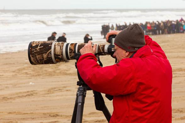 Right and in the center of the photo, a photographer in a red coat and brown wool hat looks through an enormous telephoto lens on a tripod on the beach in Scheveningen. He holds the extended telephoto lens with both hands. At the top of the photo, the sea with its waves, blurred and above the sea a narrow strip of gray sky. On the right, in the far distance, also blurred, is a long line of visitors on the beach.