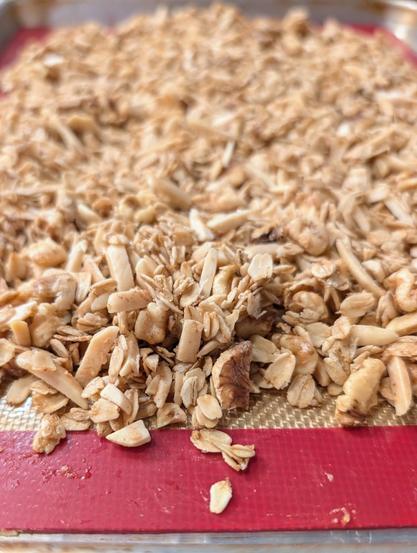 A batch of granola just out of the oven, sitting on a silicone liner inside a baking sheet.