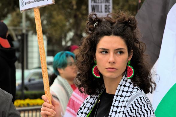 A woman is center-frame holding a placard (text not visible) and looking to the right of the camera. She appears to be serious. She is wearing a keffiyeh and large watermelon earrings.