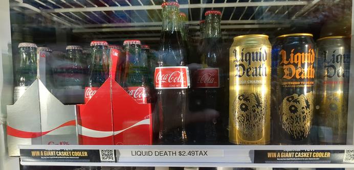 Photograph of drink refrigerator at Mr. Beet in Brooklyn. On the right are three varieties of Liquid Death. On the left are individual glass bottles of Coca Cola next to six pack glass bottles of Coca Cola and Diet Coke. There is a price sticker under the drinks which says LIQUID DEATH $2.49TAX. Curiously, the Liquid Death price sticker is under the Coca Cola instead of the Liquid Detah.