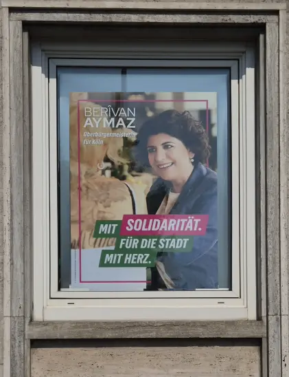 Ein modernes weißes Fenster, eingefasst von grauem Stein, darin ein Wahlkampfplakat von Berîvan Aymaz. Das Foto wurde an der Kreisgeschäftsstelle der Grünen Köln am Ebertplatz aufgenommen. Das Plakat im Fenster zeigt die Kandidatin im  Gespräch mit einer tätowierten Person mit blonden, toupierten Haaren und enthält den Text:
Berîvan Aymaz, Oberbürgermeisterin für Köln. Mit Solidarität. Für die Stadt mit Herz.