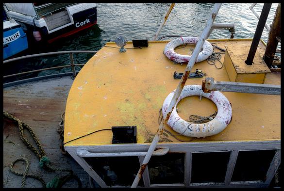 Close-up view of a yellow fishing boat deck showing life rings, rusty chains, and weathered equipment with other boats visible in Ballycotton harbour, County Cork