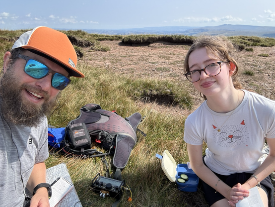 Selfie of me, with beard and orange cap. My daughter with glasses and hair tied back. Radio gear and a backpack scattered on the ground between us.